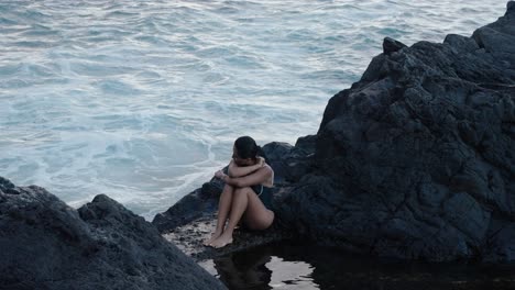A woman sits quietly on volcanic rocks by the sea in Seixal, Madeira, surrounded by crashing waves and calm tidal reflections.