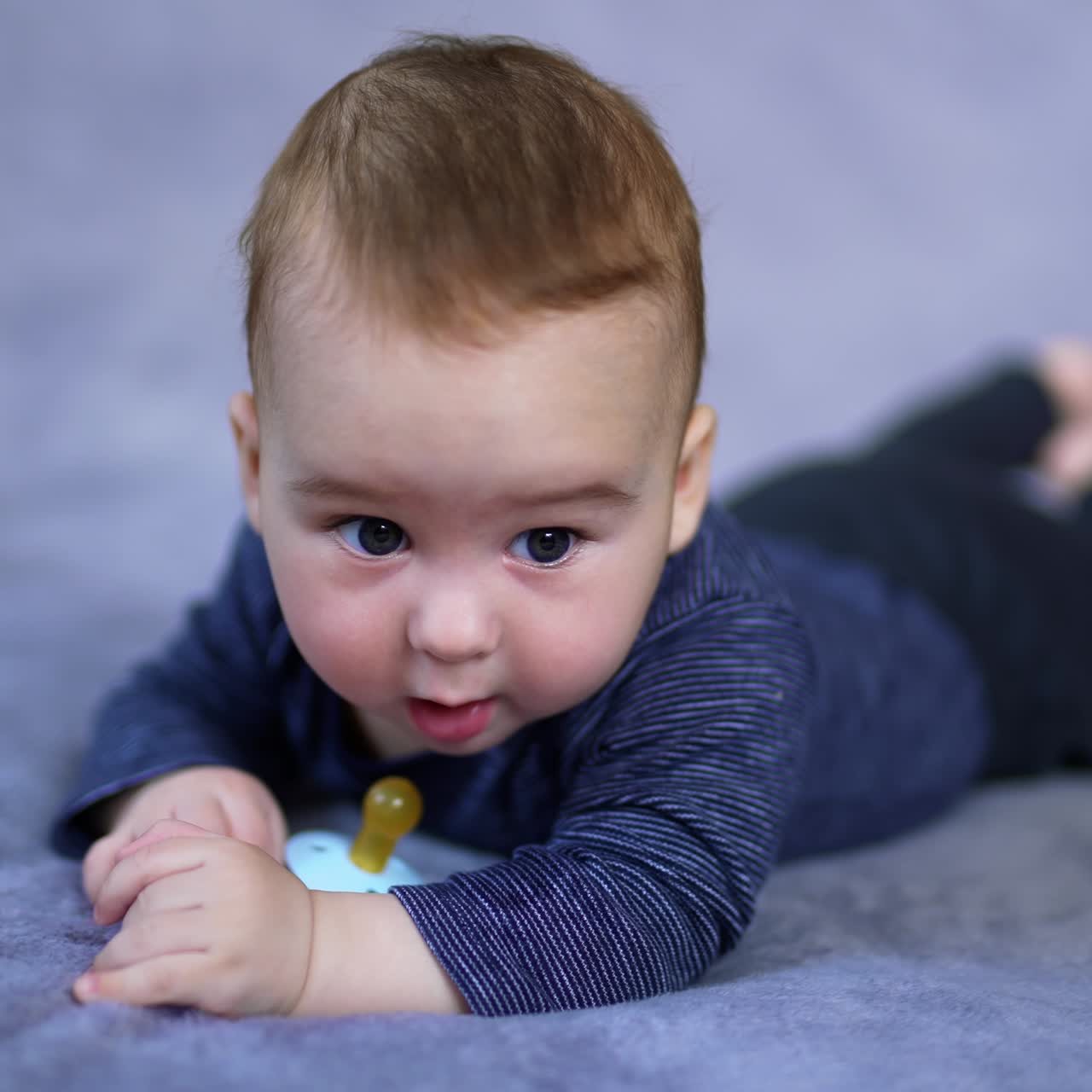 Lovely toddler in blue clothes lying peacefully on the bed. Cute child looks at pacifier with interest and then around himself