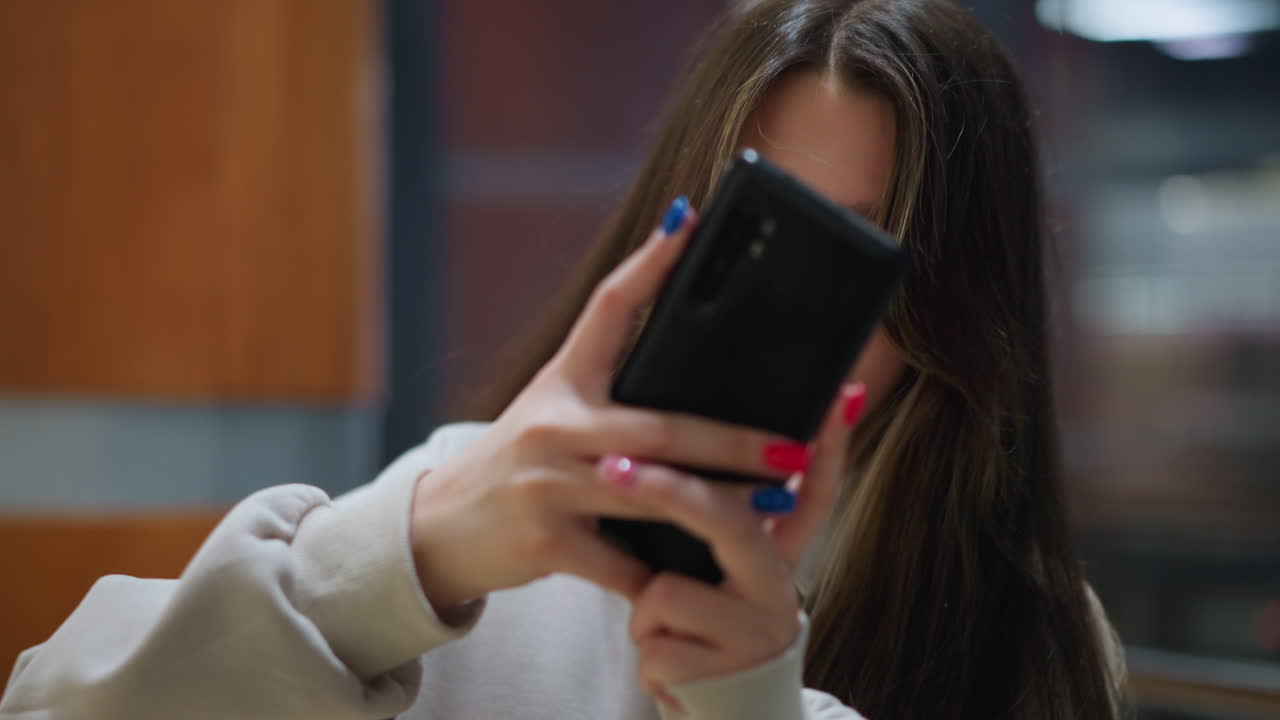 Side view of young lady concentrating while holding smartphone to take photo of person across railing indoors with evening lights reflecting on glass creating casual urban atmosphere