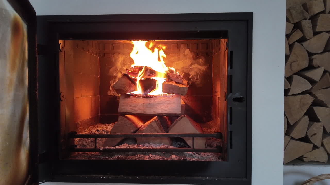A bright, steady fire burns inside a modern enclosed fireplace, with stacked firewood glowing as tall flames rise in the center. The warm orange light reflects off the fireproof interior,
