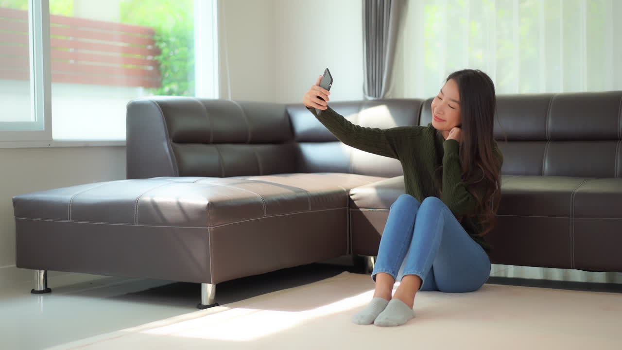 Attractive young Asian woman sit floor in home taking selfie with phone. Social media influence in lifestyle. Posing for picture on smart phone to share online. Smiling happy lady taking picture