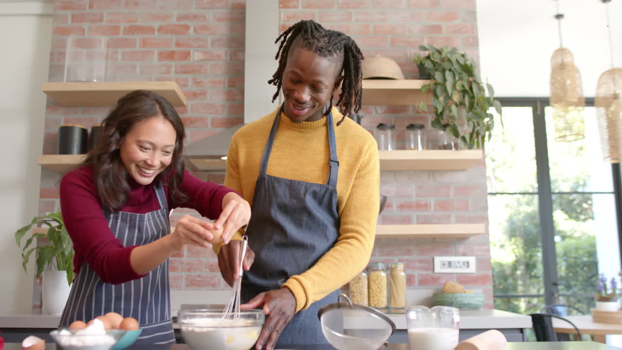 Happy diverse couple in aprons baking in sunny kitchen, copy space, slow motion