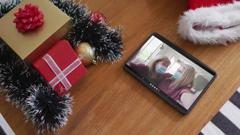una madre y una hija caucásicas sonrientes con máscaras faciales en una llamada de video de navidad en una tableta