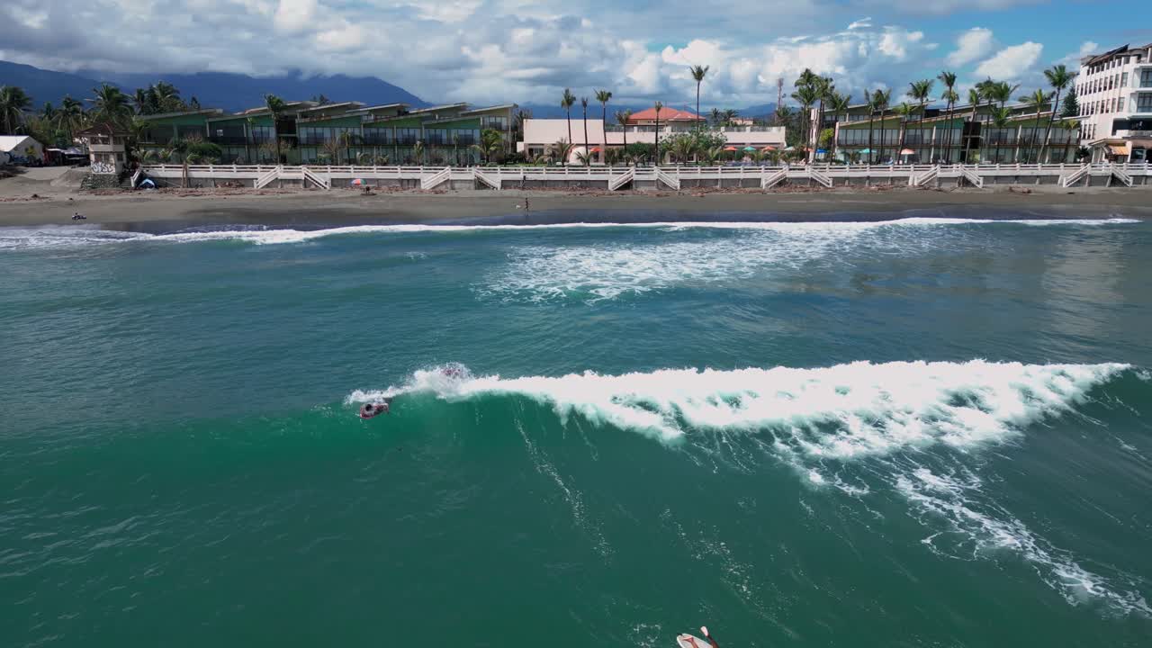 A wide drone shot captures the bustling shore, where surfers are seen riding the ocean waves, while beach houses line the coastline in the background.