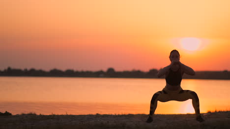 Seitenansicht-Einer-Jungen-Frau,-Die-Draußen-Kniebeugen-Macht.-Seitenansicht-Einer-Jungen-Fitness-Frau,-Die-Kniebeugen-Macht-Und-Bei-Sonnenuntergang-In-Zeitlupe-Am-See-Auf-Dem-Sand-Steht