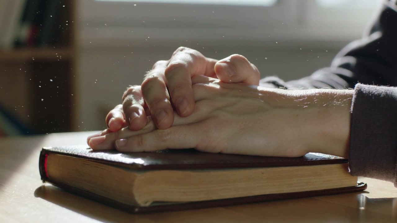 Mani appoggiate con maniche scure, giunte su un libro rilegato chiuso vicino a una finestra illuminata dal sole, con particelle di polvere che fluttuano.