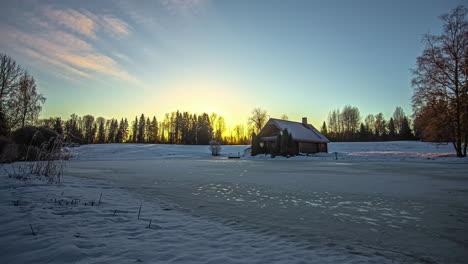 Pequeña-Cabaña-En-La-Nieve-Bajo-Nubes-Naranjas-En-Un-Cielo-Dorado-En-Un-Paisaje-Nevado-Con-árboles-En-Un-Lapso-De-Tiempo-De-Invierno