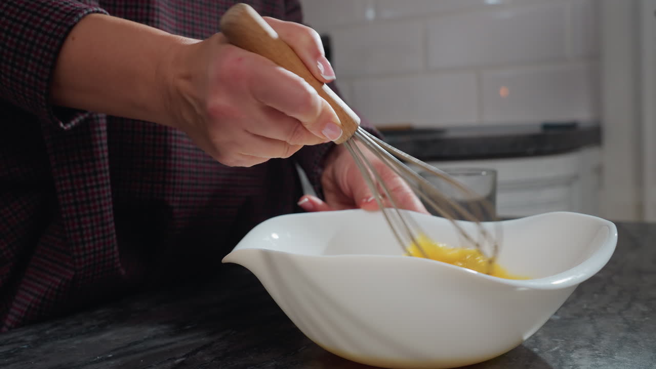 vista lateral de primer plano del chef golpeando huevos en un cuenco blanco en la encimera oscura, la mano mezcla firmemente las yemas, creando una textura suave para hornear, batedor con mango de madera en movimiento, taza de vidrio vacía cerca