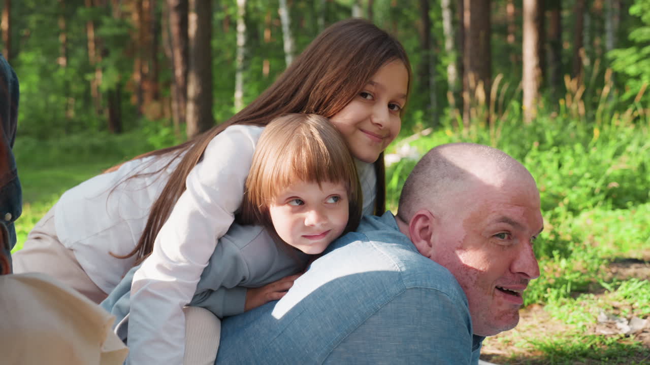 Primo piano di due ragazze che si riposano sulla schiena del papà all'aperto sotto la calda luce del sole, circondate dalla famiglia in una tranquilla ambientazione nella foresta, che mostra amore, legame affettivo e affetto durante una giornata felice e serena insieme nella natura
