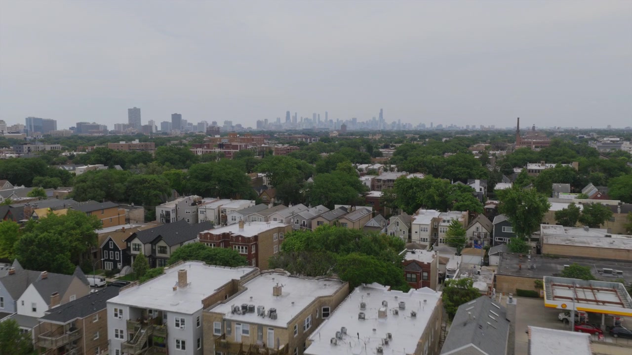 Vista aérea de drone sobre um bairro arborizado de Chicago com telhados e torres de igrejas, com o horizonte do centro da cidade visível ao longe, envolto em neblina