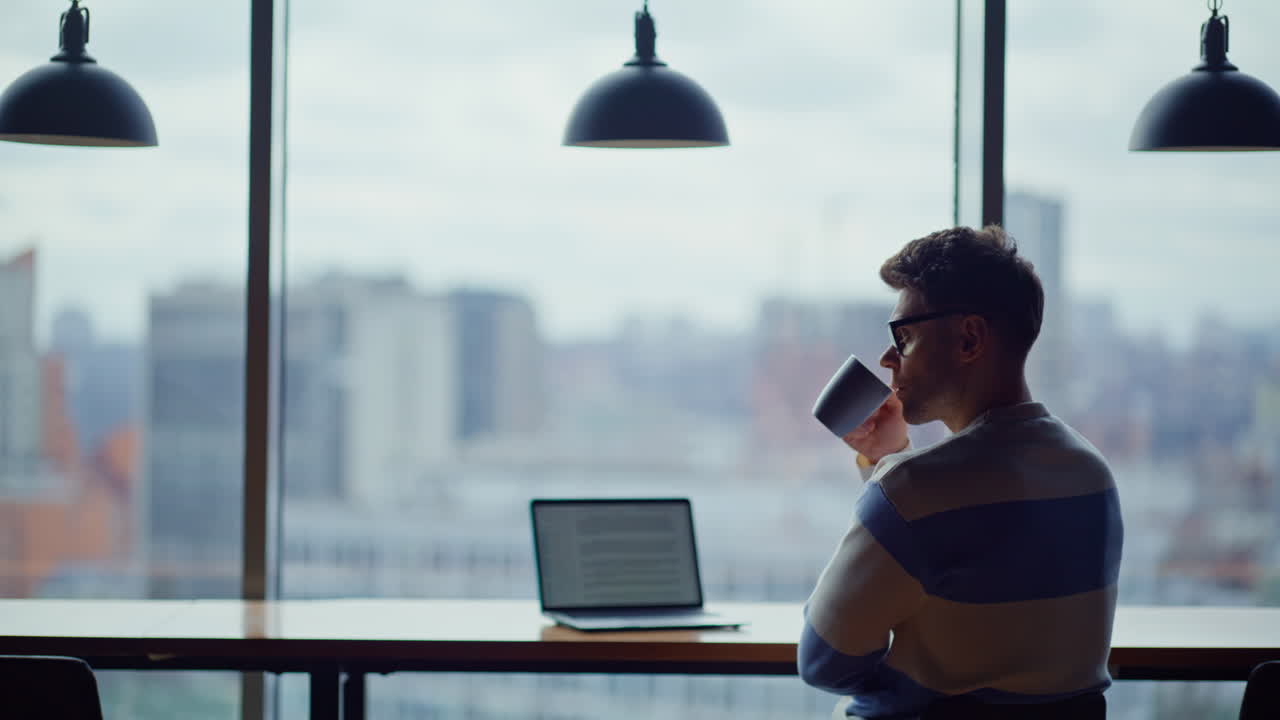 Afslappet programmør, der drikker te og kigger ud i det fjerne i et panorama-kontor med udsigt over byen
