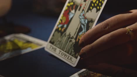 Close-Up-Of-Woman-Giving-Tarot-Card-Reading-On-Candlelit-Table-Holding-The-Tower-Card-2