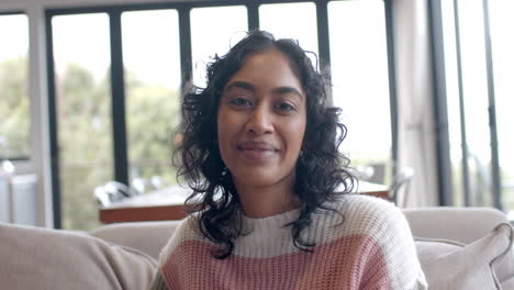 portrait of happy biracial woman sitting on sofa smiling at home, slow motion