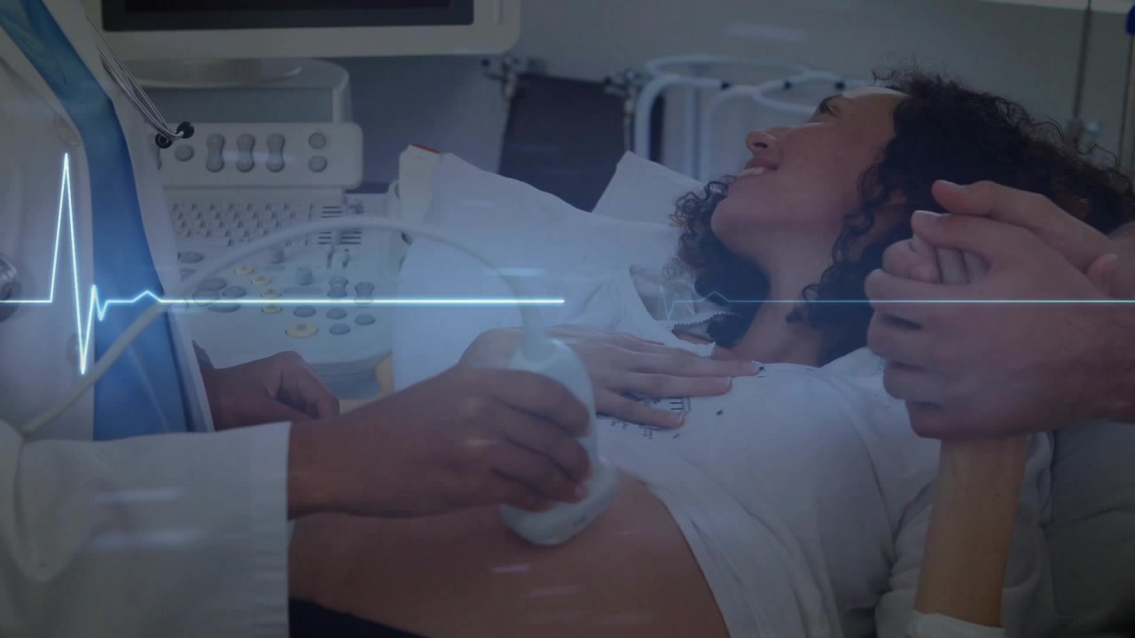 Ecografista che applica il gel e passa la sonda sull'addome, con il partner che tiene la mano per il monitoraggio ECG fetale.