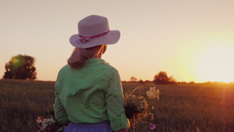 a romantic woman with a hat and a bouquet of wildflowers admires the sunset over the wheat field