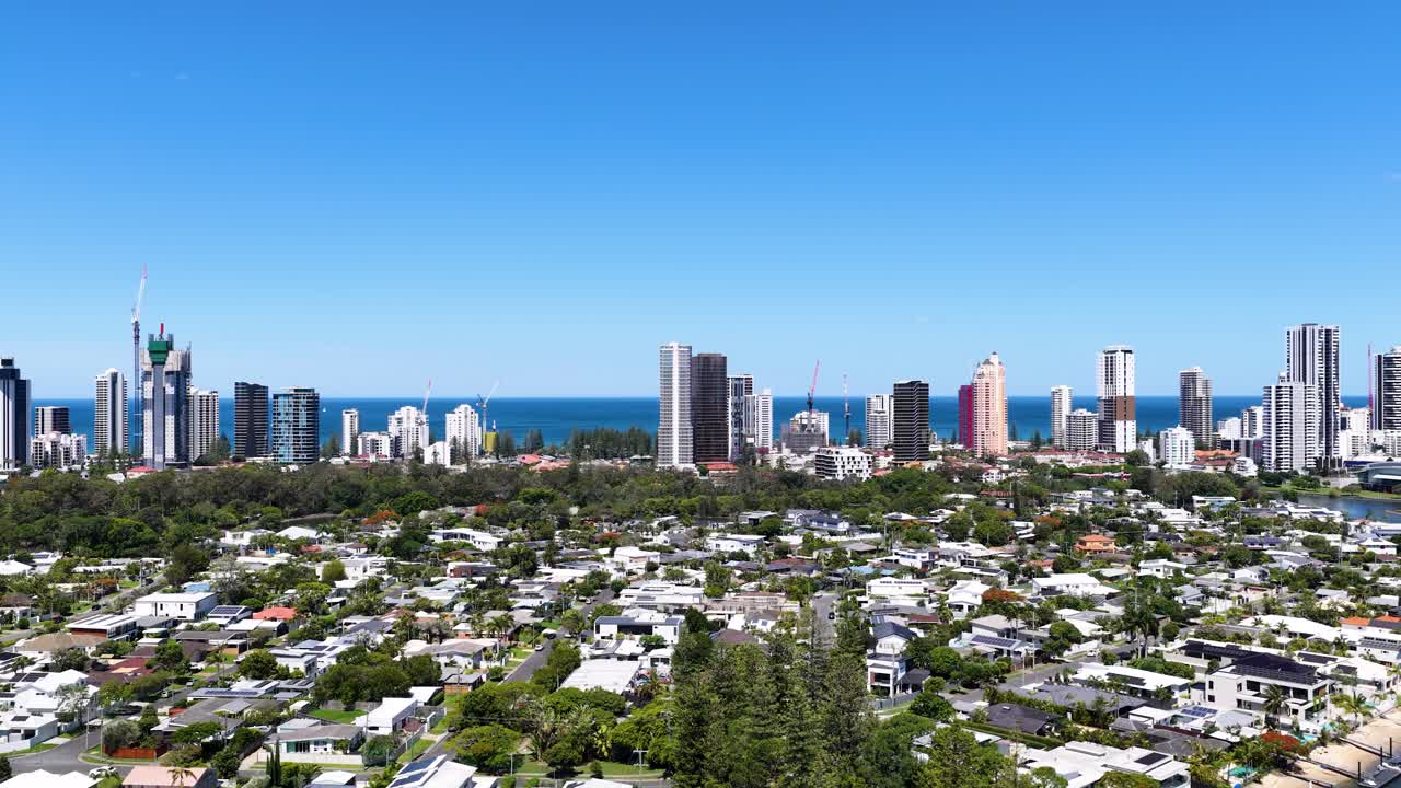 Drone ascends above residential canal, revealing Gold Coast skyline, ocean horizon, and clear blue sky