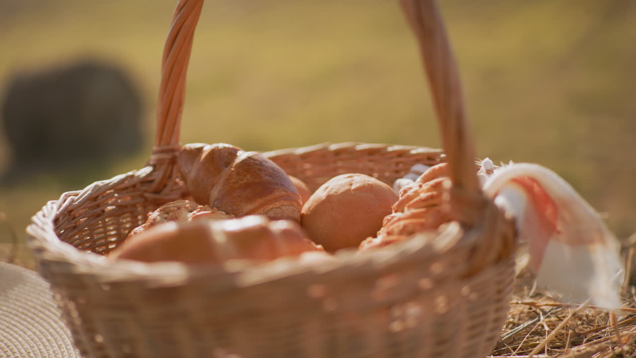 Close-up of fresh golden croissants and bread rolls in woven picnic basket with napkin, set against blurred open field under warm sunlight