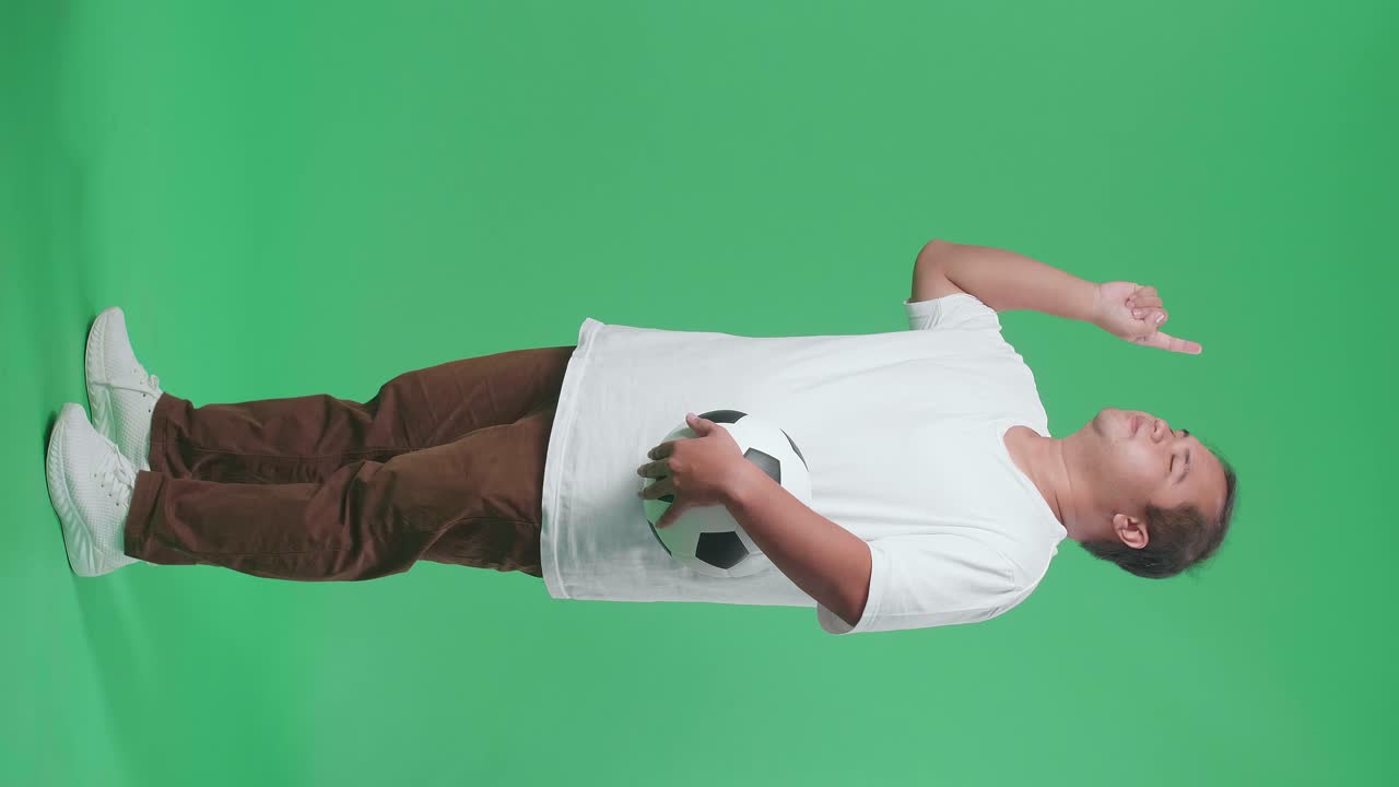 눈이 닫힌 몸으로 가득 찬 아시아 남자가 공을 들고 지표손가락을 올리고 머리를 흔들며 녹색 화면 배경에서 축구를 응원합니다.