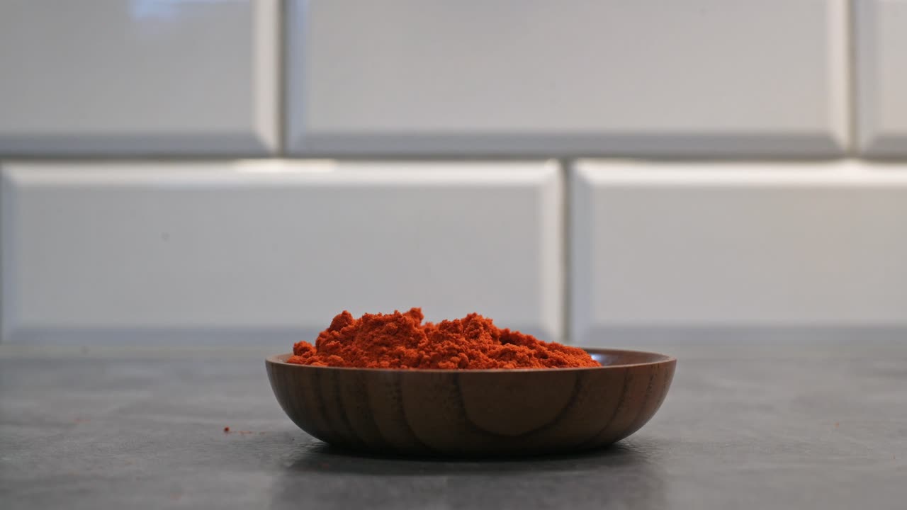 A close-up of ground paprika in a wooden bowl with a small wooden scoop, held by a hand. The video highlights the vibrant red color of the spice, ideal for culinary themes.