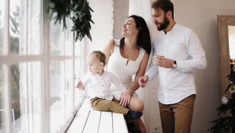 Junge-Schöne-Familie-Sitzt-Zusammen-Am-Fenster,-Geschmückt-Mit-Weihnachtskranz.-Liebevolle-Eltern,-Die-Sich-Gegenseitig-Küssen