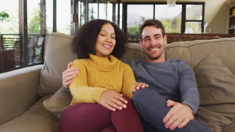 Portrait-of-happy-diverse-couple-sitting-on-couch-in-living-room,-embracing-and-laughing