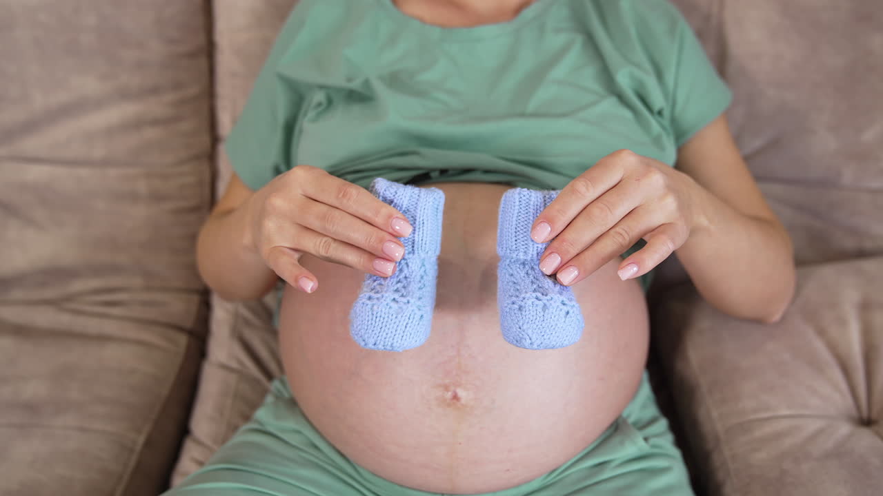 Pregnant, beautiful woman sitting on the sofa with little blue baby booties. Woman moves the tiny socks back and forth on her big bare belly.
