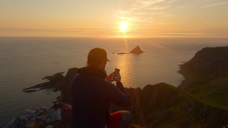 Men-taking-a-picture-with-his-phone-of-the-sunset-at-the-ocean-and-island-Bleiksoya-rock-landscape-in-Norway