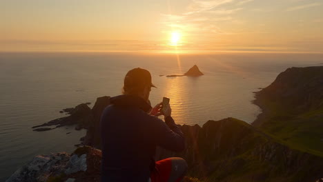 Hombres-Tomando-Una-Foto-Con-Su-Teléfono-De-La-Puesta-De-Sol-En-El-Océano-Y-El-Paisaje-Rocoso-De-La-Isla-Bleiksoya-En-Noruega