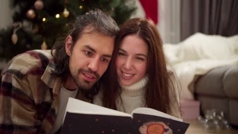 an interested guy in a checkered shirt and a brunette girl in a white sweater with a cup of hot drink in their hands are reading a new year's book in the cozy atmosphere of a room decorated in new year's style