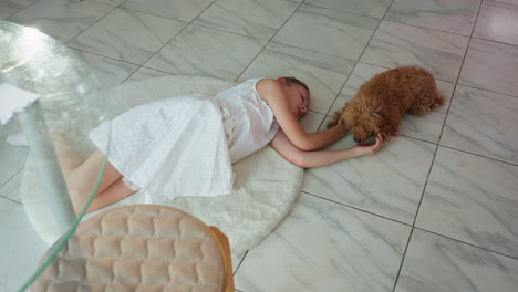 little girl in white gown lying on white rug, smiling as her dog licks her hand. background features a chair and glass table, capturing peaceful indoor bonding moments between child and pet