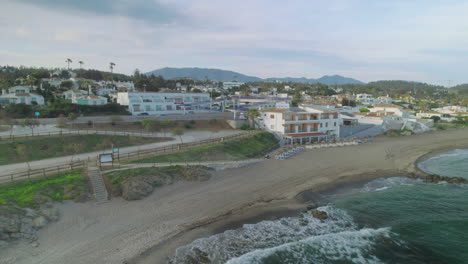 Antena-Moviéndose-Diagonalmente-Hacia-Atrás-Cerca-De-La-Playa-De-Mijas-Costa,-Andalucía,-España,-En-Un-Día-Nublado-Y-Cerca-De-Las-Carreteras-Costeras