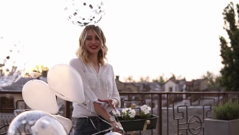 mujeres jóvenes rubias caucásicas con blusa blanca y vaqueros afuera en la terraza o balcón. girando con globos blancos transparentes. disfruta de su tiempo. felicidad. paisaje de la ciudad en el fondo