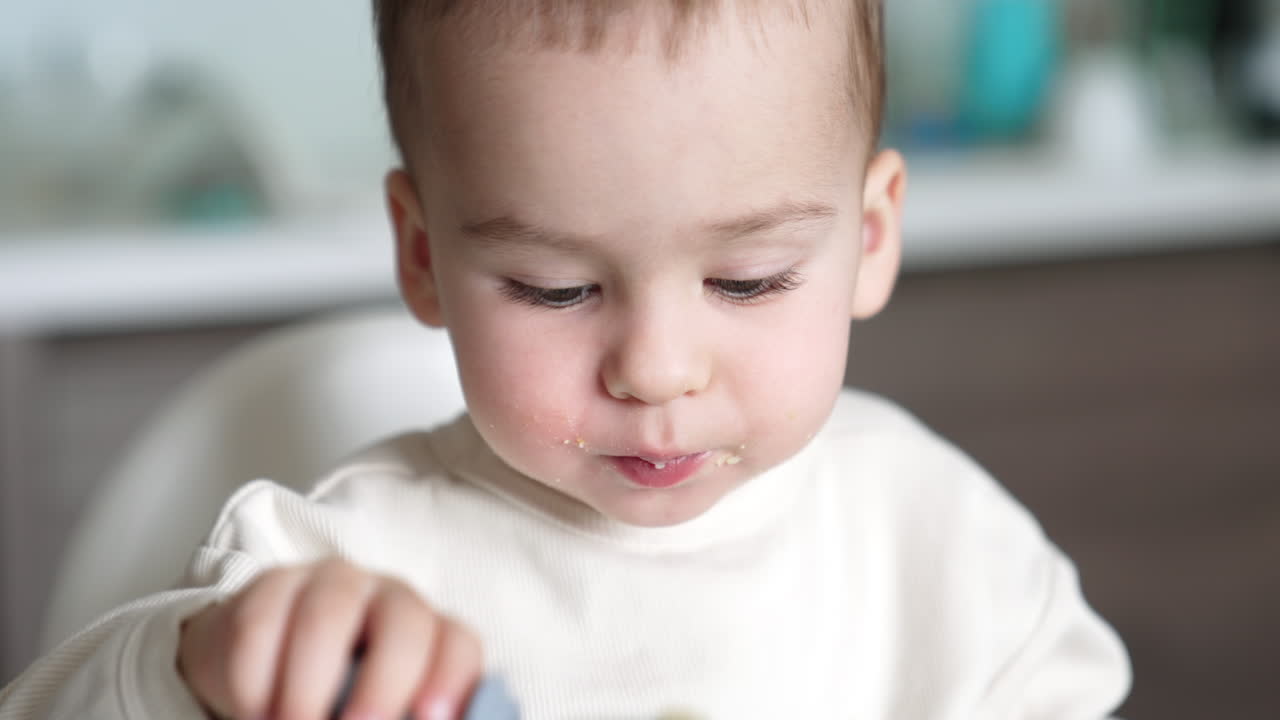 Lovely Caucasian baby boy with pieces of food on his face. Little child eating from spoon himself. Close up.
