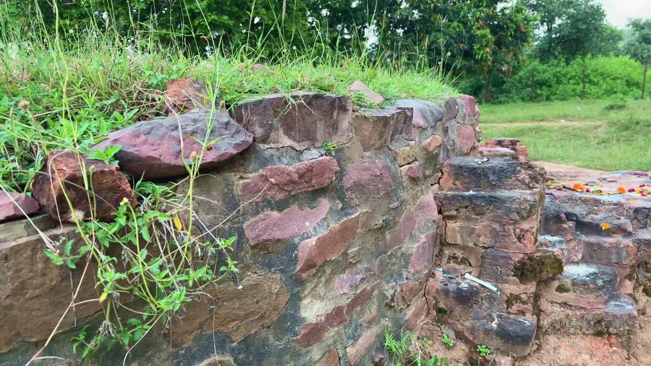 A left-to-right tracking shot of the weathered brick and stone wall at the Bharhut Stupa site in Madhya Pradesh, showing centuries-old craftsmanship now covered with offerings and moss