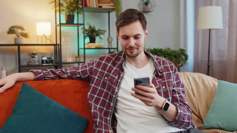 Un-Joven-Feliz-Usando-Un-Teléfono-Móvil-Viendo-Mensajes-De-Texto-De-Películas-En-Línea-En-Un-Teléfono-Inteligente-En-La-Habitación-De-Casa