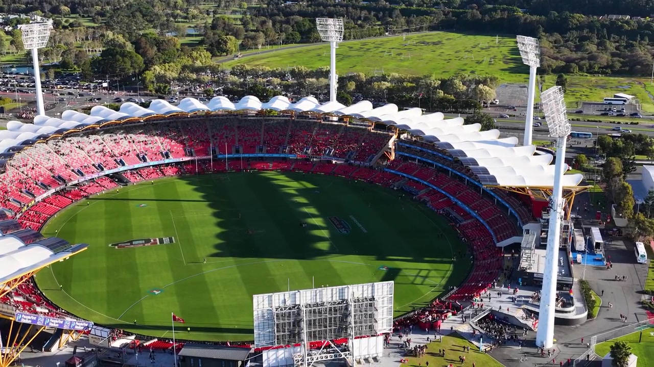 High-angle drone shot pans across a circular sports arena filled with spectators under sunlight.