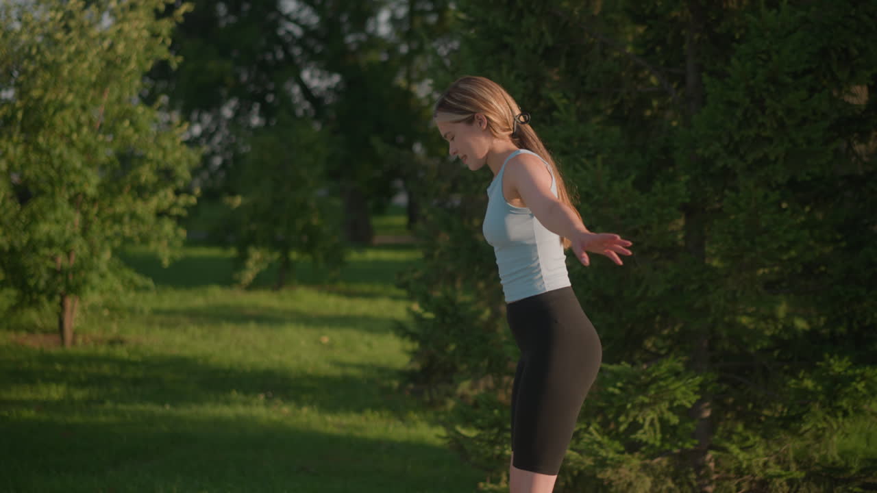 Side view of young white woman looking down while skating outdoors, swinging her hands for balance on sunny day, surrounded by lush greenery and trees, near grassy field