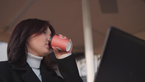 woman in black coat, sitting under patio, takes a sip of her tea cup, bursts into laughter, and gently places the cup back on table, a partial laptop is in front of her