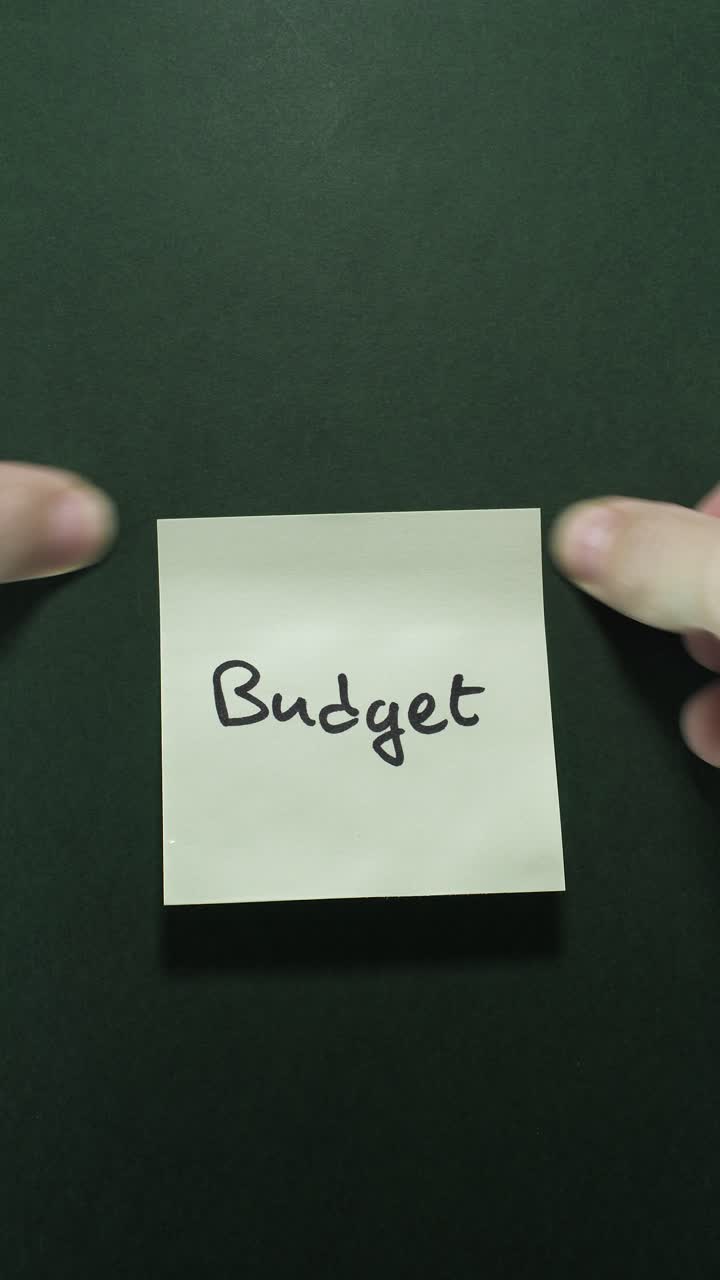 Hænder sætter en "Budget"-post-it op på en mørkegrøn væg, der udforsker temaet personlig økonomi.