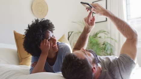 Video-De-Una-Feliz-Pareja-Diversa-Relajándose-En-Casa,-Acostada-En-La-Cama-Usando-Un-Teléfono-Inteligente