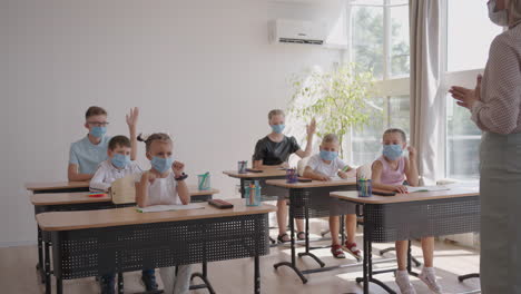 levanten la mano para responder a la pregunta del maestro. grupo multiétnico de niños con máscaras faciales en la escuela durante la pandemia de covid-19.