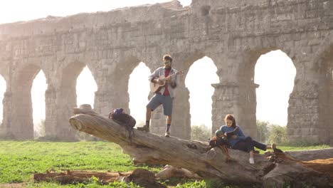 happy young couple backpackers tourists on a log trunk playing guitar singing reading book in front of ancient roman aqueduct ruins in romantic parco degli acquedotti park in rome at sunrise slow motion