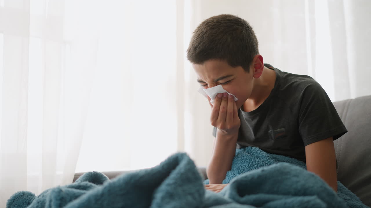 Sick boy sneezing into tissue seated on couch with blanket covering him, looking tired near window with glass cup on table, showing signs of fatigue while managing cold symptoms