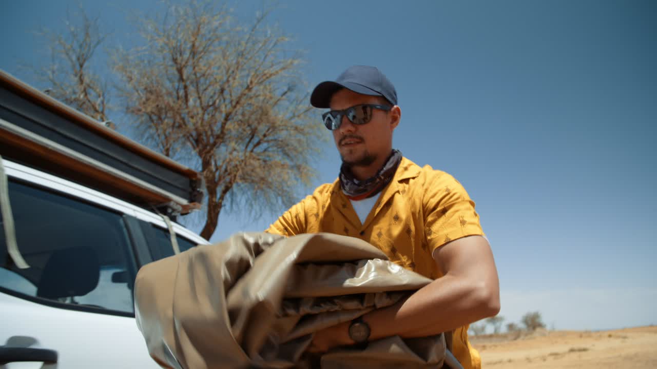 Handheld tracking shot of a tourist folding and putting a rooftop tent cover in the truck of his off-road vehicle in Africa.