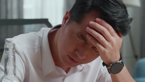 close up of drunk asian man with vodka glass holding the temples while sitting on the chair at home