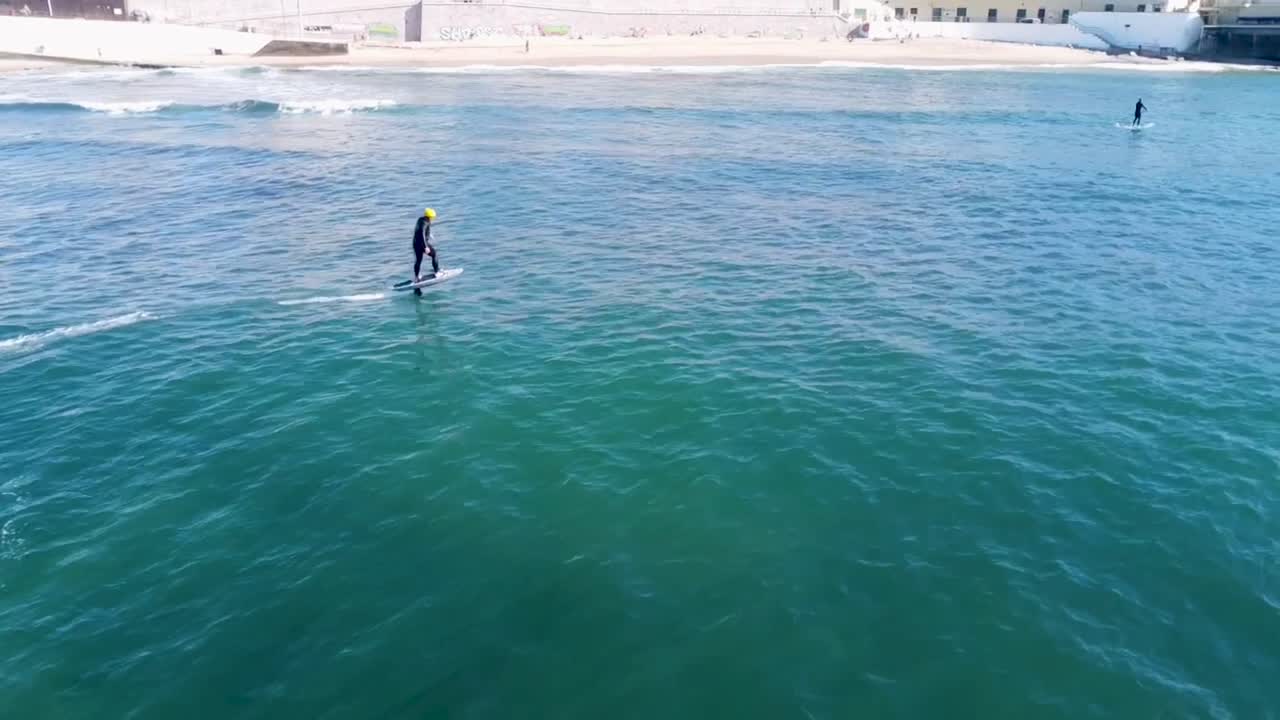 Athlete on a hydrofoil surfboard riding a wave, using a foil drive assist pumps to catch a wave on the outside.