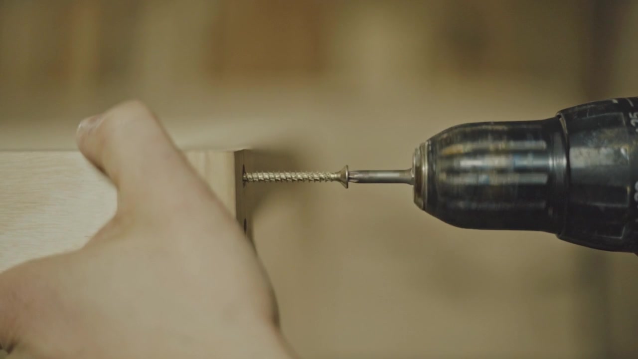 Close up of craftsman driving screw into wooden board with power drill, fastening timber during woodworking project on workbench inside carpentry workshop
