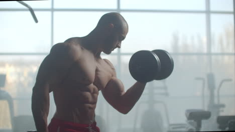 a man with a naked torso and a pumped strong body and relief muscles of the press raises dumbbells against the background of the window training his hands. strength of will and stubborn character.