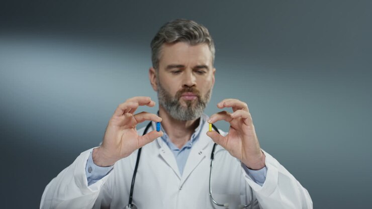 Male Nice Cute Doctor Holding Two Pills In Hands Yellow And Blue And Looking At Them Like Comparing And Choosing Blue One
