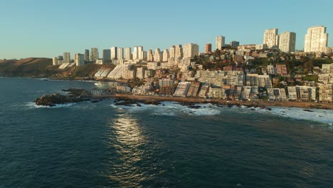 Aerial-view-flying-across-Reñaca-sunny-cityscape-beach-coastline-reflecting-in-ocean